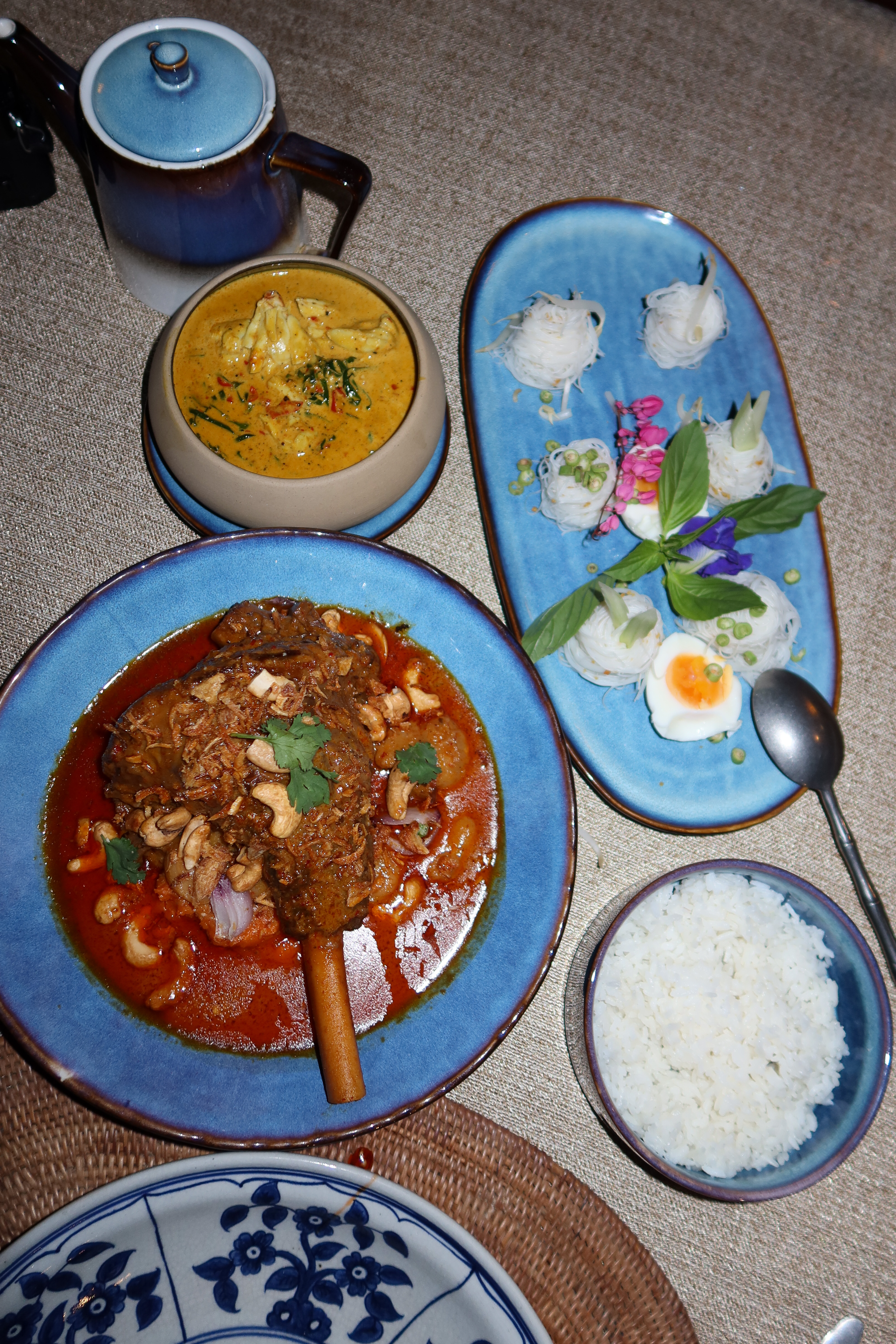 Elegant Thai dinner spread at Krua Phranang in Rayavadee, featuring rich crab curry, lamb shank massaman curry, jasmine rice, and delicate rice noodle nests with herbs and a soft-boiled egg.