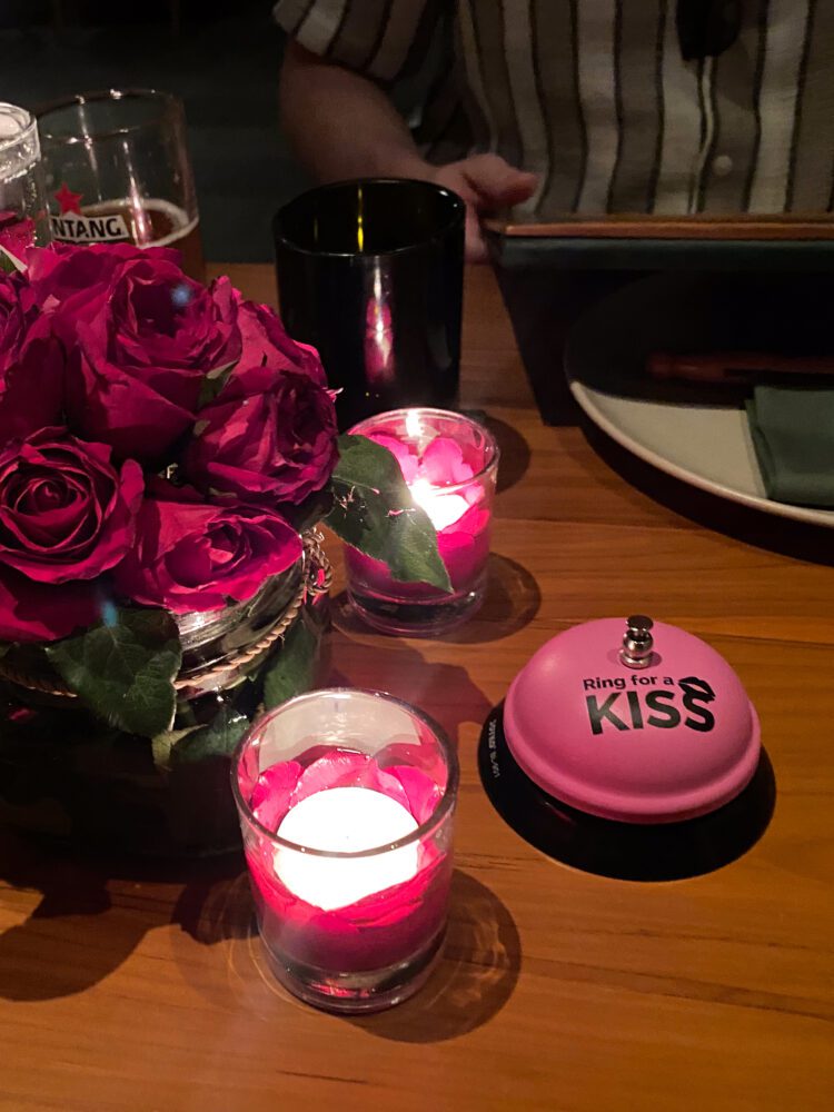 Candlelit dinner table at Cuca Uluwatu with red roses, glowing votives, and a playful pink “Ring for a Kiss” bell.