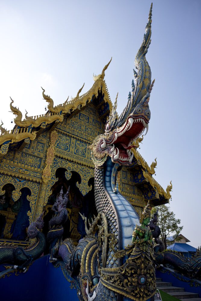 Intricately detailed dragon statue guarding the entrance to Wat Rong Suea Ten (Blue Temple) in Chiang Rai, Thailand, with vibrant blue and gold temple architecture against a clear sky.