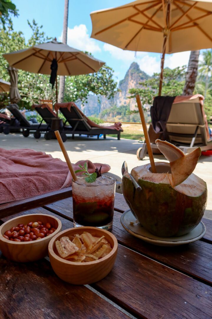 Tropical poolside refreshments at Rayavadee’s Pool Bar, featuring a fresh coconut, a fruit-infused cocktail, and local snacks with a backdrop of loungers and dramatic limestone cliffs.