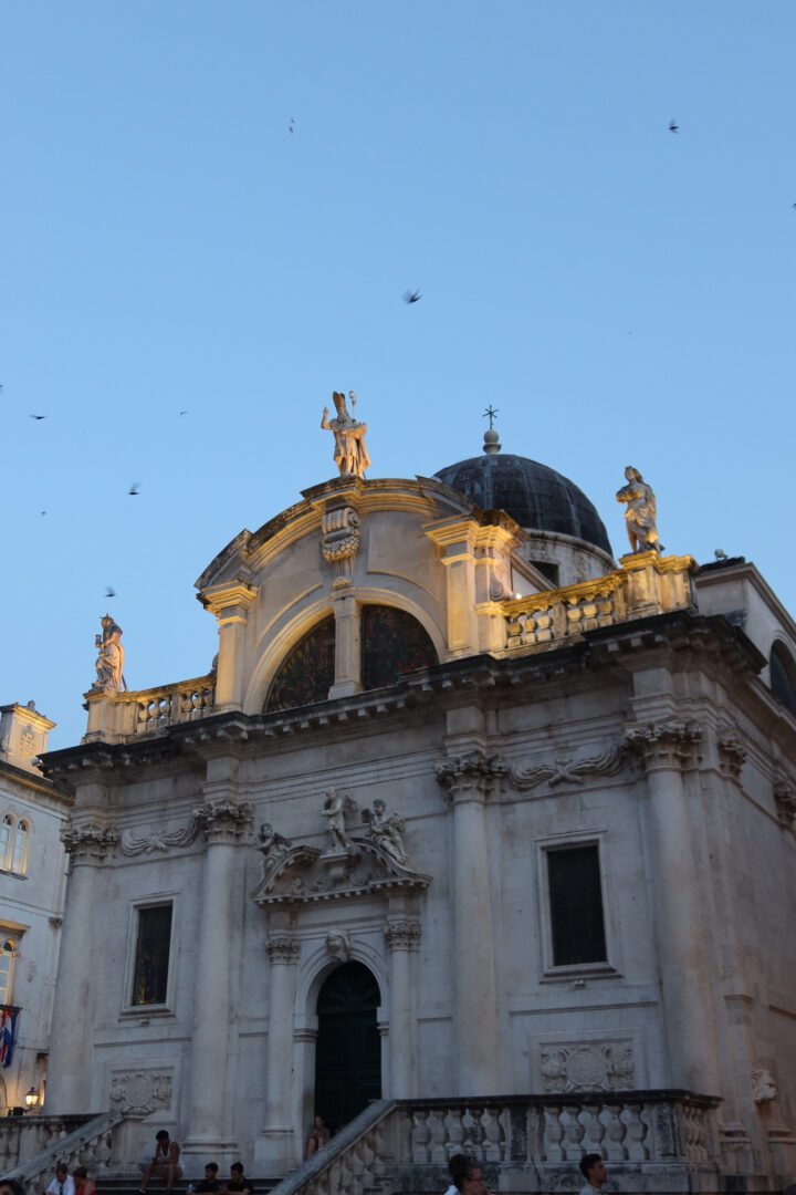 Dubrovnik Cathedral glowing at dusk, with statues and baroque details illuminated against the evening sky.