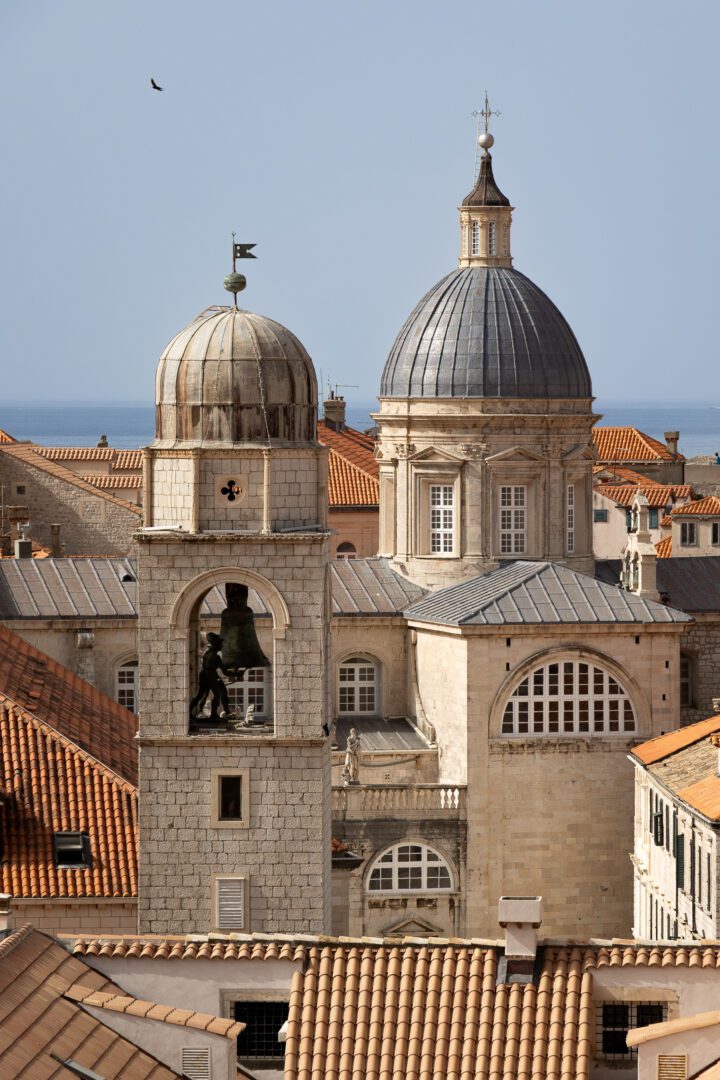 Scenic view of Dubrovnik Cathedral and bell tower rising above terracotta rooftops with the Adriatic Sea in the background.