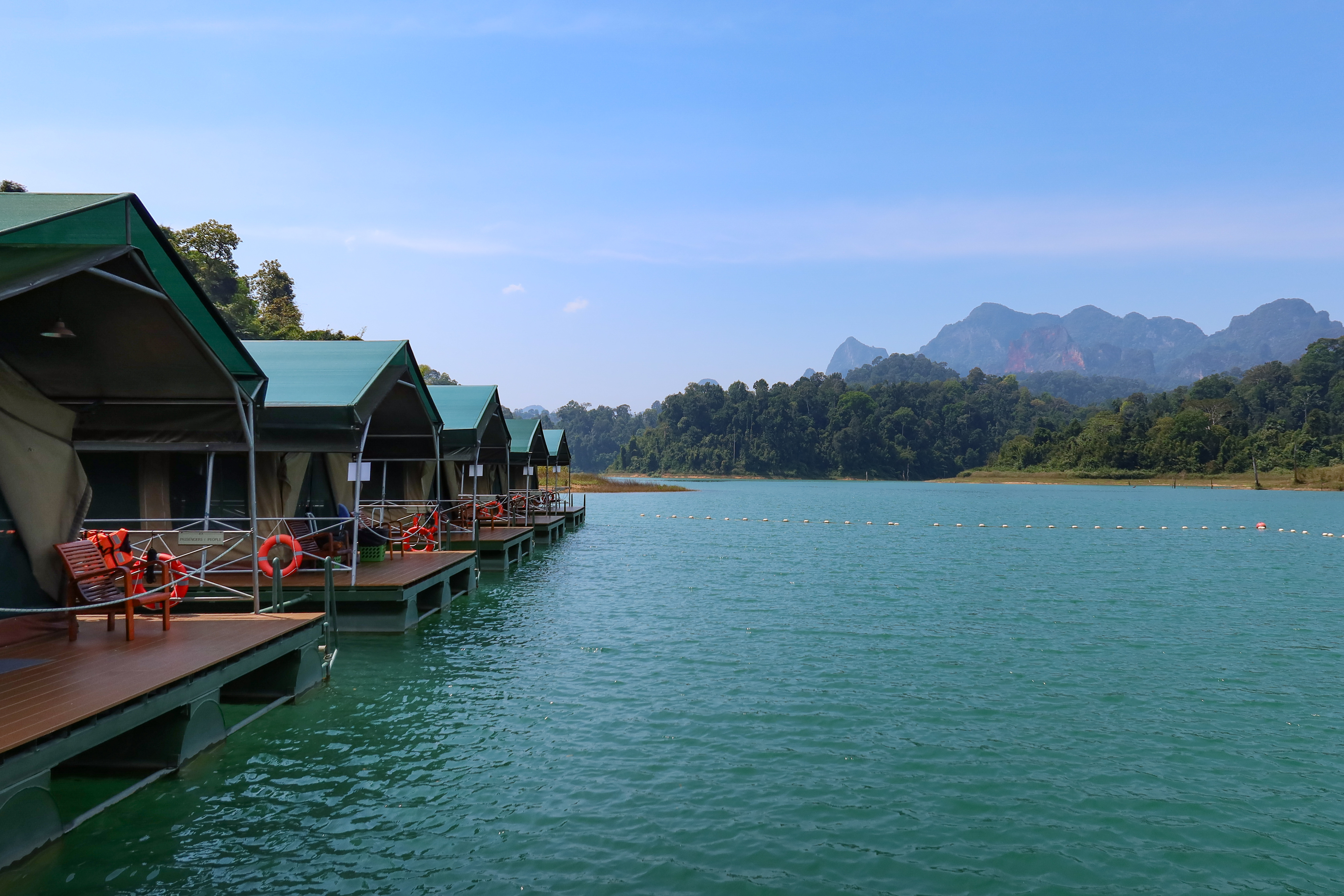 A row of floating tents at Elephant Hills Lake Camp stretches over the emerald waters of Cheow Lan Lake, with jungle-covered mountains rising in the distance under a clear blue sky.