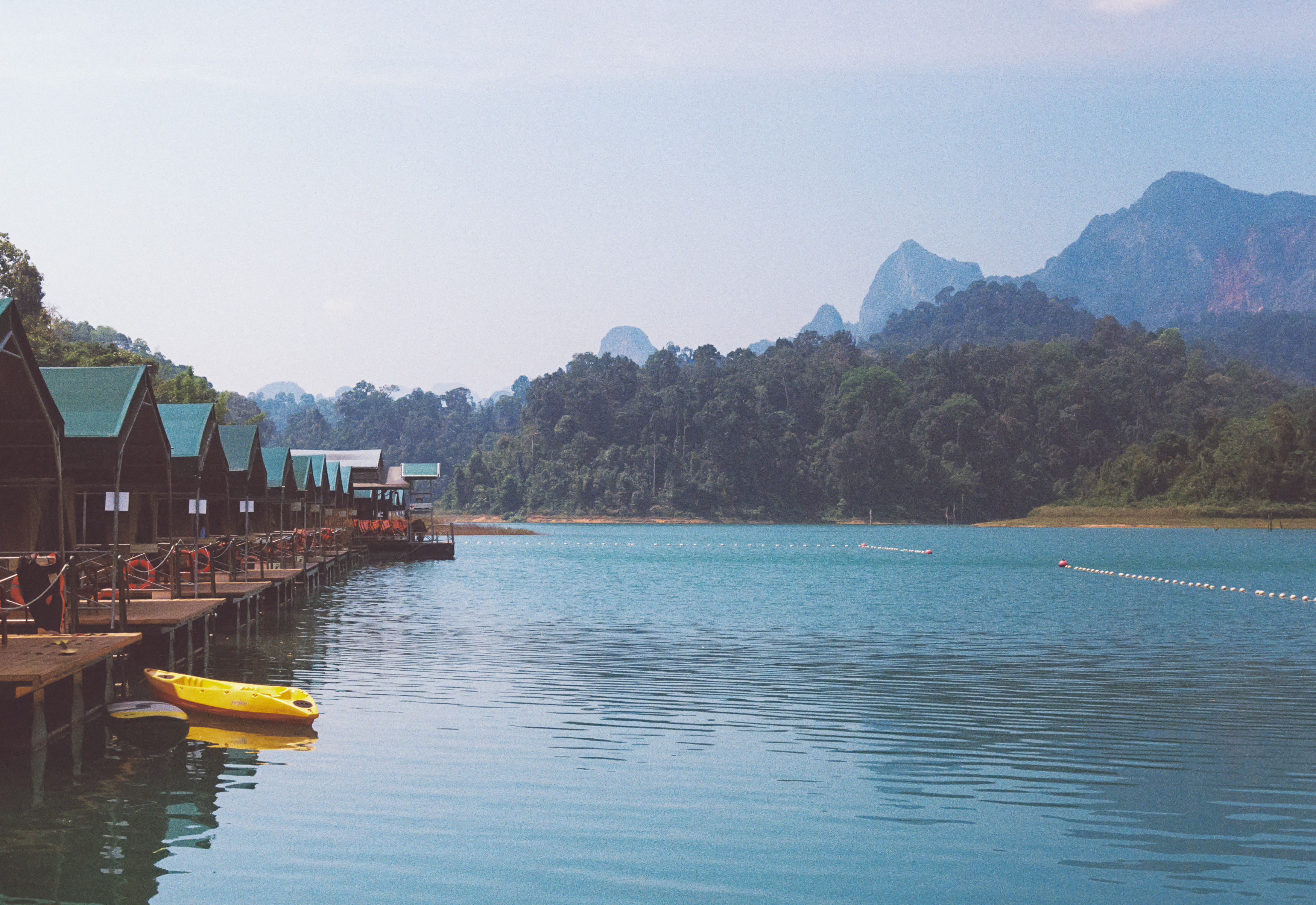 A row of floating tents lines the tranquil turquoise waters of Cheow Lan Lake, with jungle-covered mountains rising in the hazy distance at Elephant Hills Lake Camp.