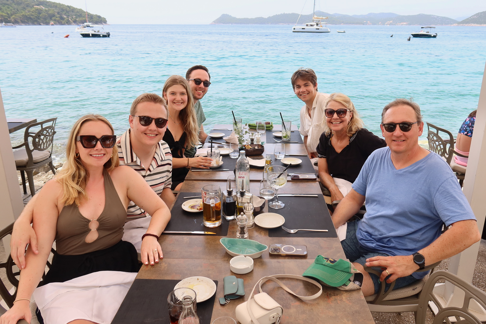Family enjoying lunch together at a seaside restaurant on Lopud Island with turquoise waters and sailboats nearby.