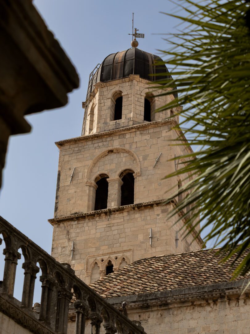 Bell tower of the Franciscan Monastery framed by palm fronds and stone architecture in Dubrovnik’s Old Town.