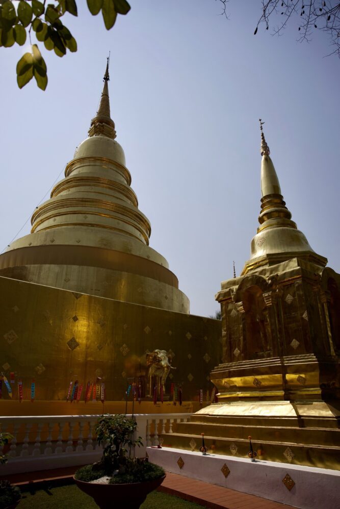 Golden stupa at Wat Phra Singh in Chiang Mai, Thailand, gleaming under the midday sun with intricate gold detailing, flanked by smaller chedis and traditional decorative banners in a peaceful temple courtyard.