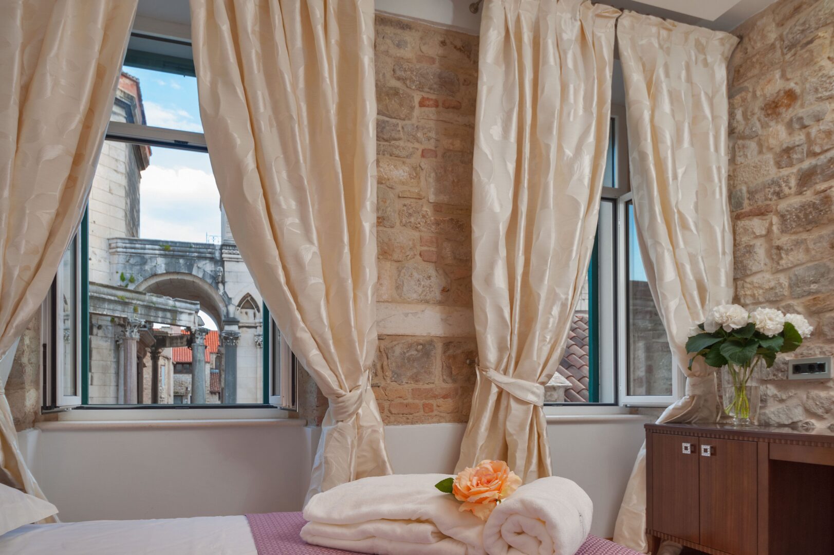 Charming hotel room in Split, Croatia with exposed stone walls and a view of Diocletian’s Palace through elegant cream-colored curtains.