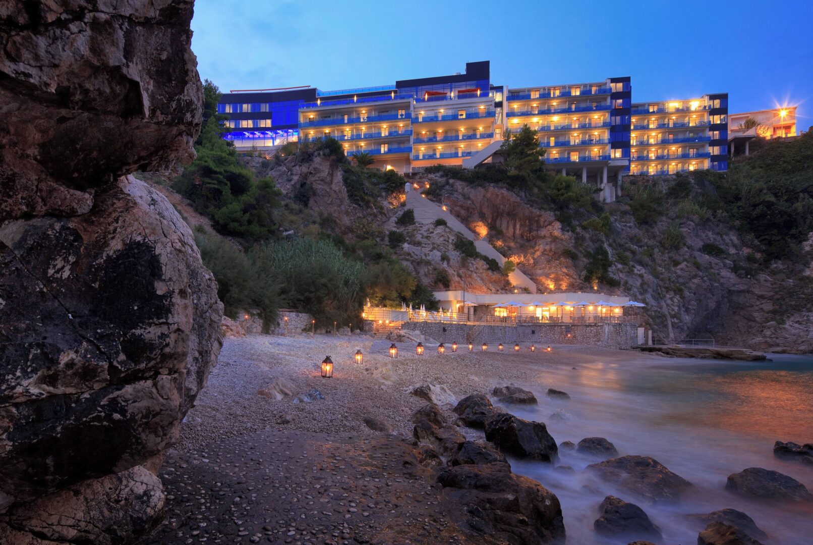Hotel Bellevue Dubrovnik illuminated at dusk, perched above a secluded pebble beach with glowing lanterns lining the shoreline and dramatic cliffs rising on either side.