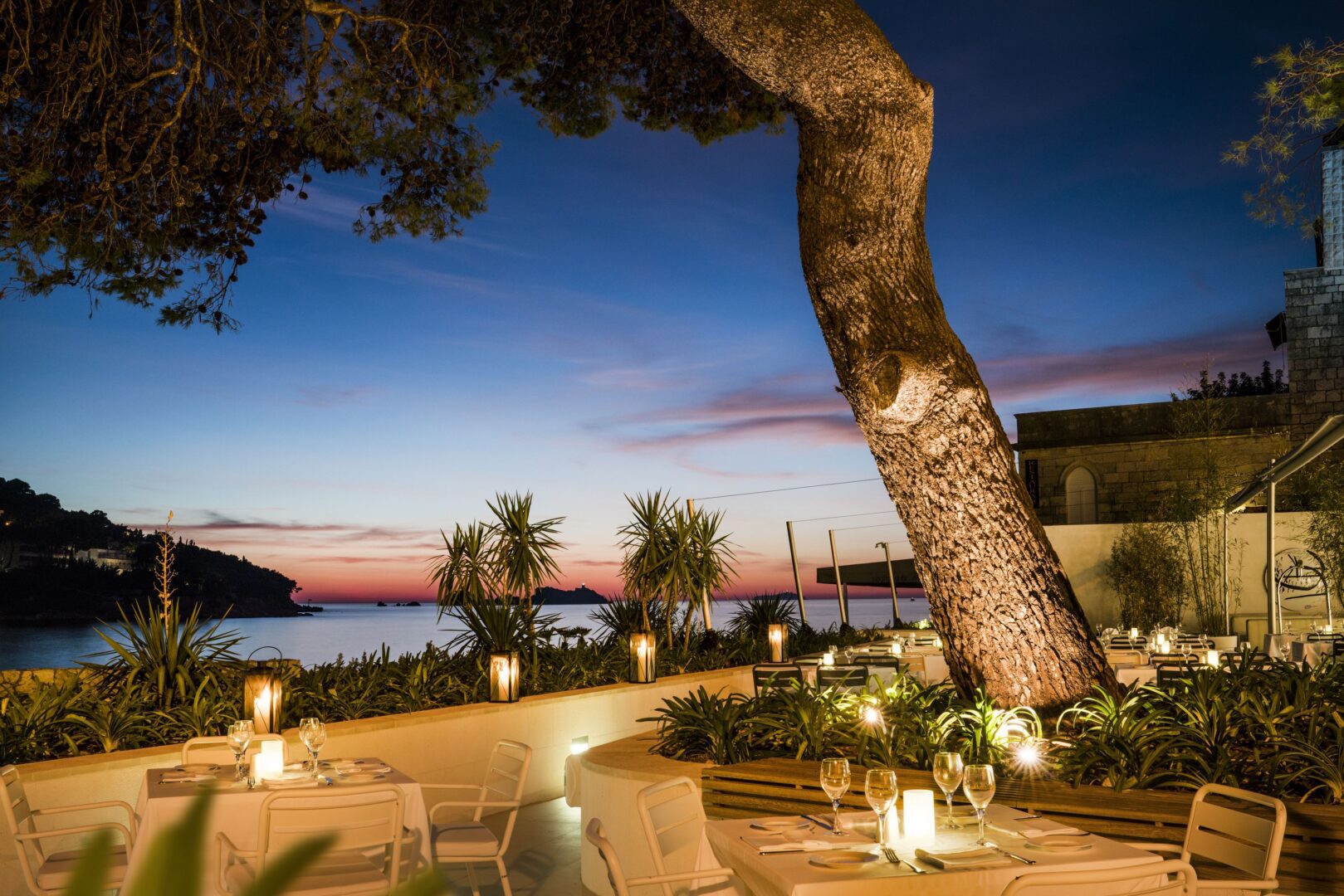 Romantic outdoor dining at Hotel Kompas Dubrovnik, with candlelit tables, lush greenery, and a serene Adriatic sunset casting warm hues over Lapad Bay.