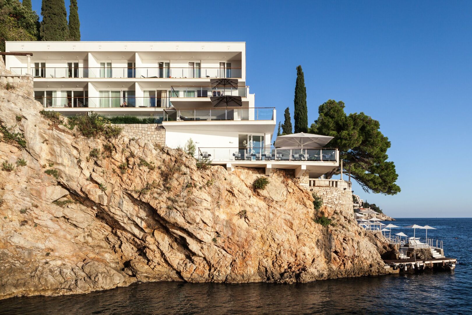 Modern white hotel perched dramatically on a rocky cliffside above the Adriatic Sea in Dubrovnik, Croatia, with private terraces and a chic waterfront lounge area overlooking the water.
