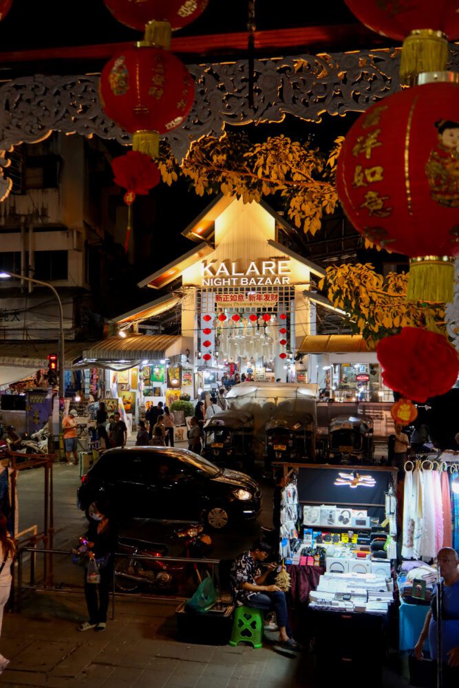 Entrance to Kalare Night Bazaar in Chiang Mai, Thailand, lit up at night with red lanterns and festive decorations during Chinese New Year, surrounded by tuk-tuks, market stalls, and crowds of visitors.