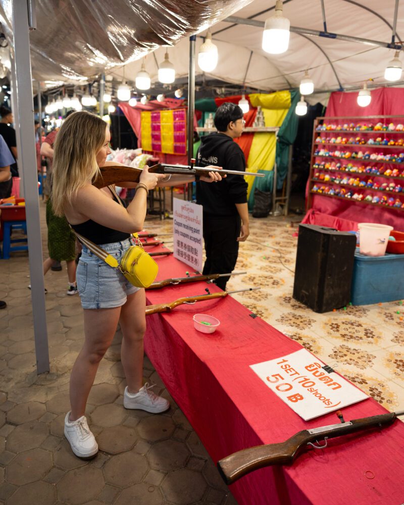 Tourist aiming a pellet rifle at a carnival-style shooting game booth inside Kalare Night Bazaar in Chiang Mai, Thailand, surrounded by colorful lights, prize displays, and playful market atmosphere.