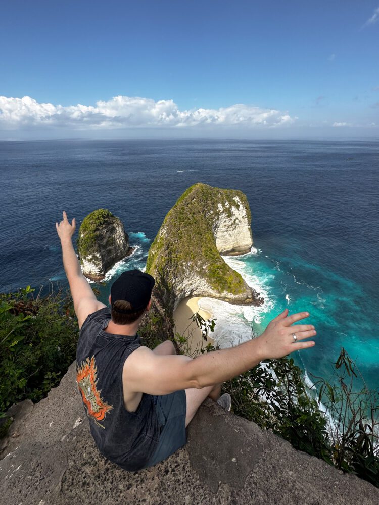 Man sitting at the edge of the cliff at Kelingking Beach, Nusa Penida, with arms outstretched toward the iconic T-Rex-shaped rock formation and turquoise waters below.