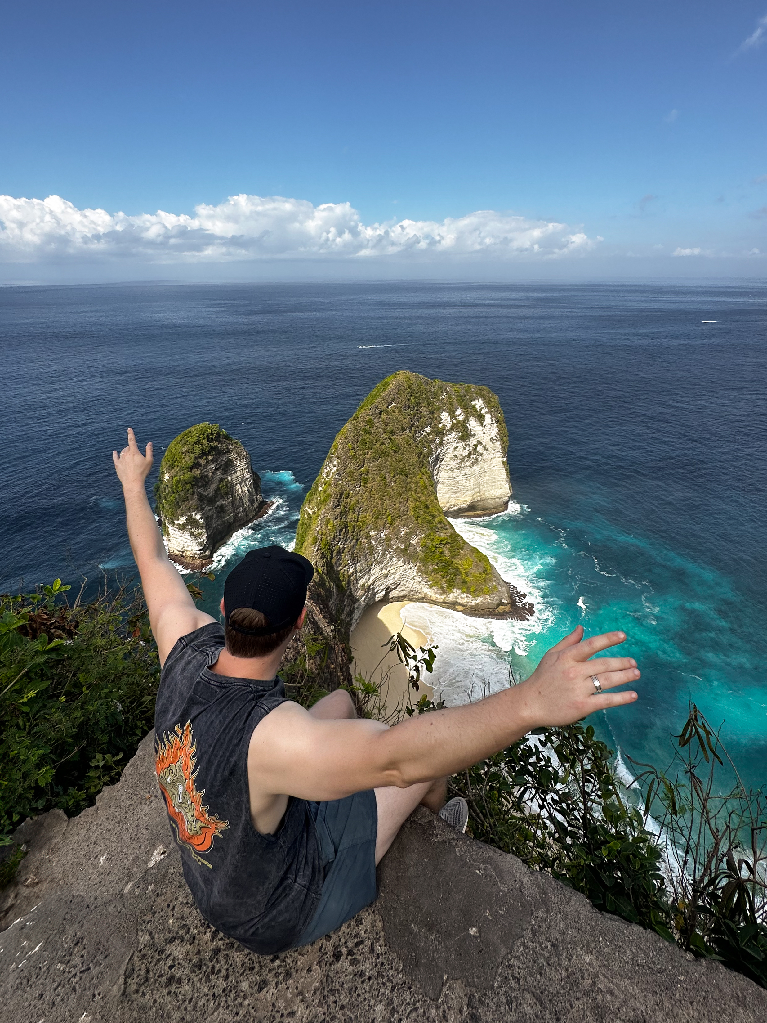Man sitting at the edge of the cliff at Kelingking Beach, Nusa Penida, with arms outstretched toward the iconic T-Rex-shaped rock formation and turquoise waters below.
