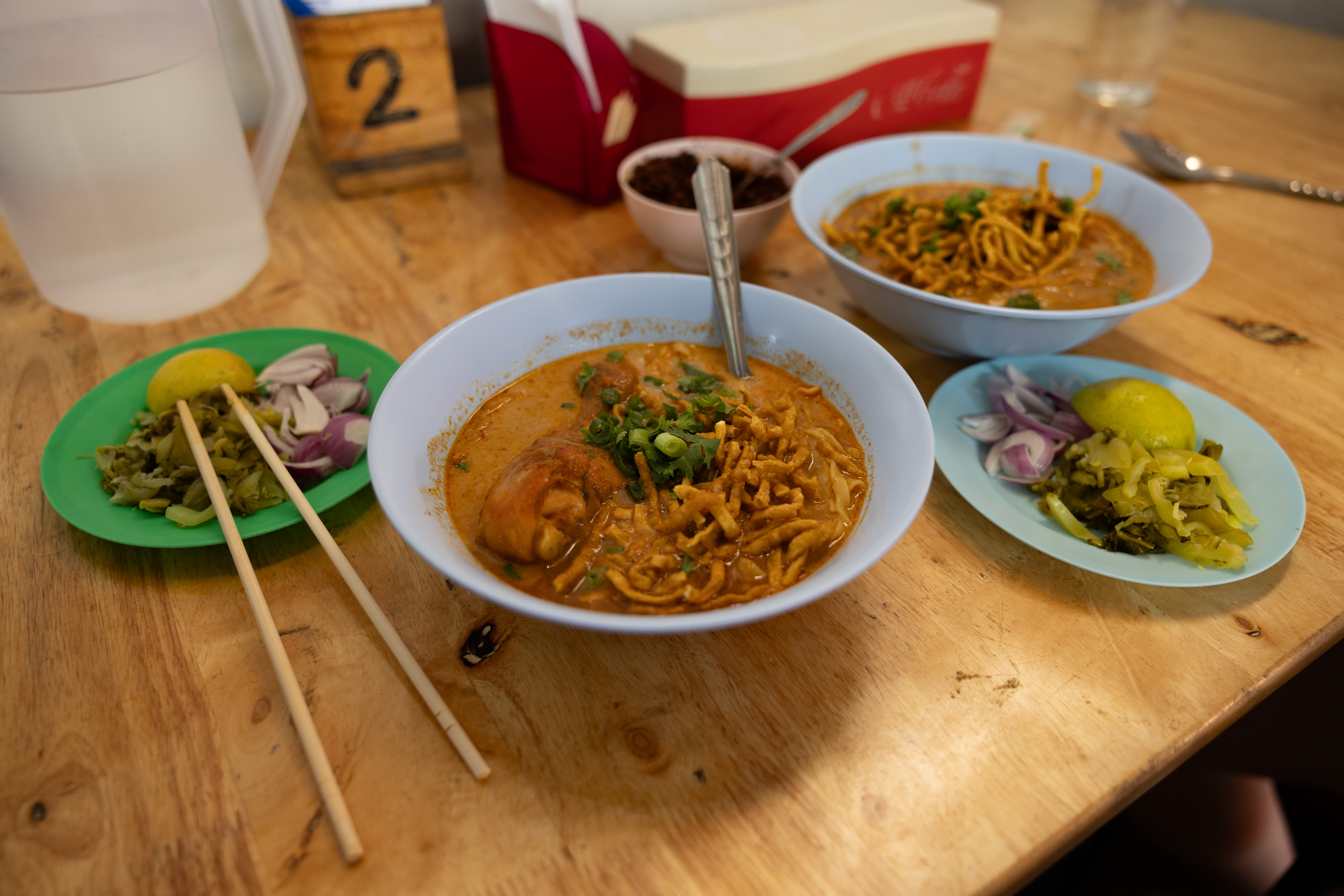 Traditional bowls of khao soi served at Khao Soi Lung Prakit Kad Kom in Chiang Mai, Thailand, featuring rich curry broth, crispy noodles, and chicken, accompanied by lime, pickled mustard greens, and shallots.