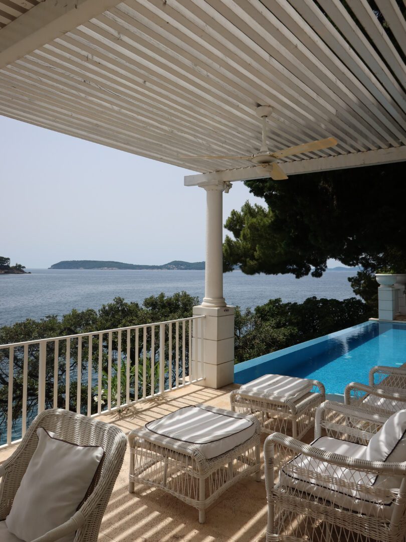 Shaded terrace with white wicker lounge chairs overlooking a serene infinity pool and the Adriatic coastline near Dubrovnik.