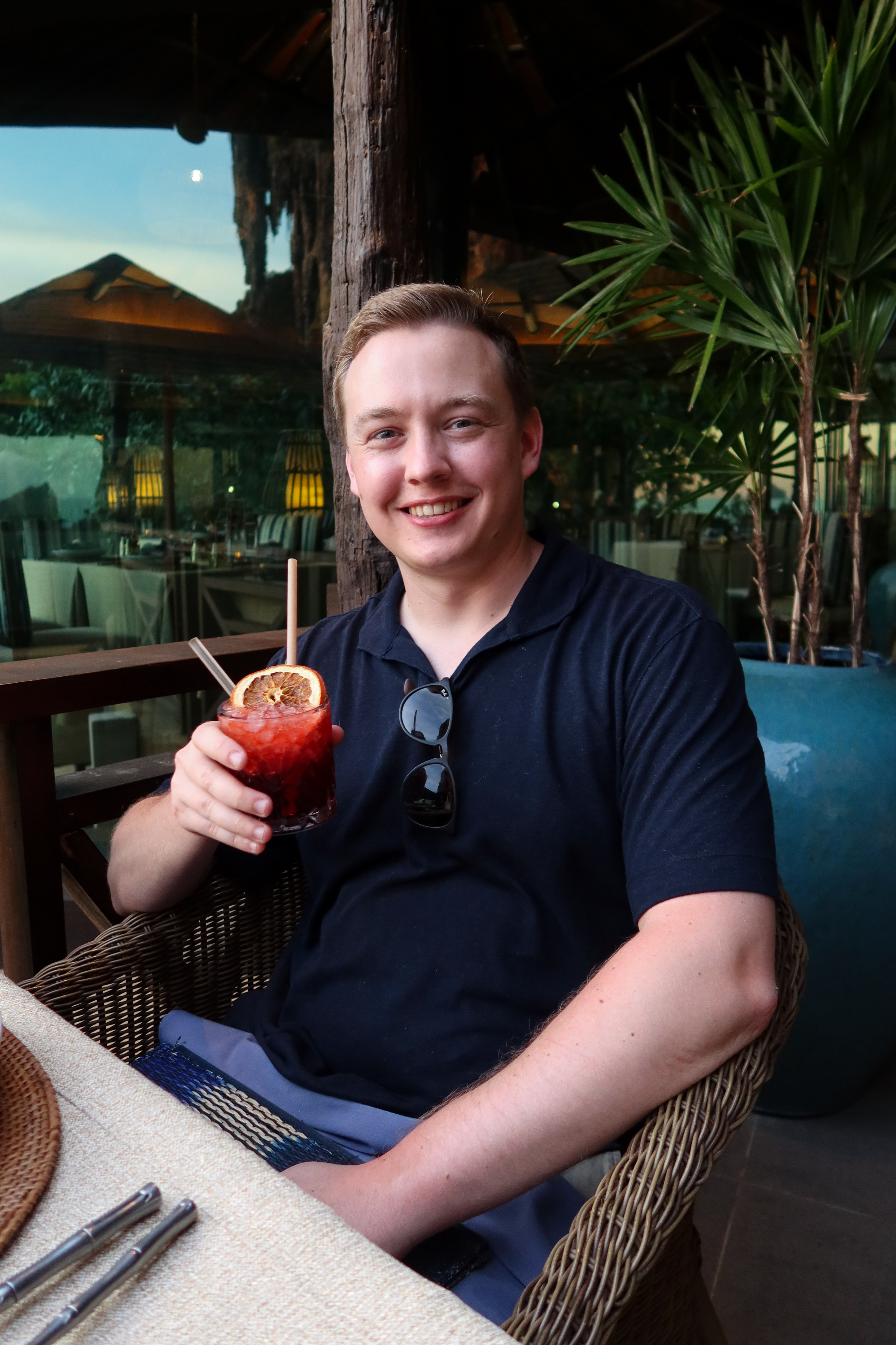 Smiling guest enjoys a handcrafted cocktail garnished with dried citrus while dining al fresco at Krua Phranang, Rayavadee’s upscale Thai restaurant nestled in a lush, tropical setting.