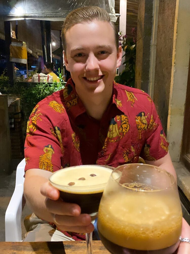 Man in a red tiger-print shirt smiling and holding up two espresso martinis while dining at a Bali restaurant.