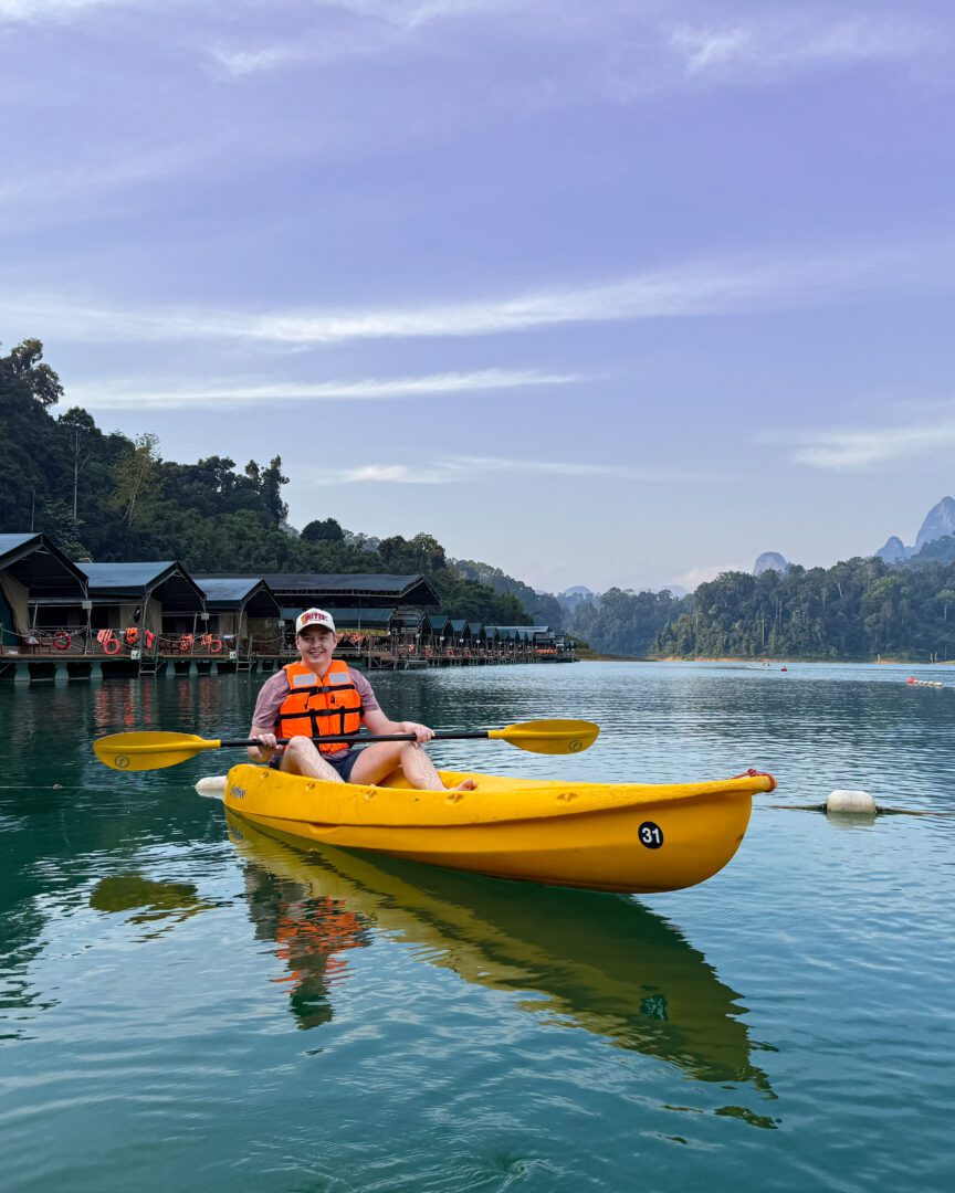 A man paddles a bright yellow kayak on the calm, turquoise waters of Cheow Lan Lake, with floating tents of Elephant Hills Lake Camp and misty mountains in the background.