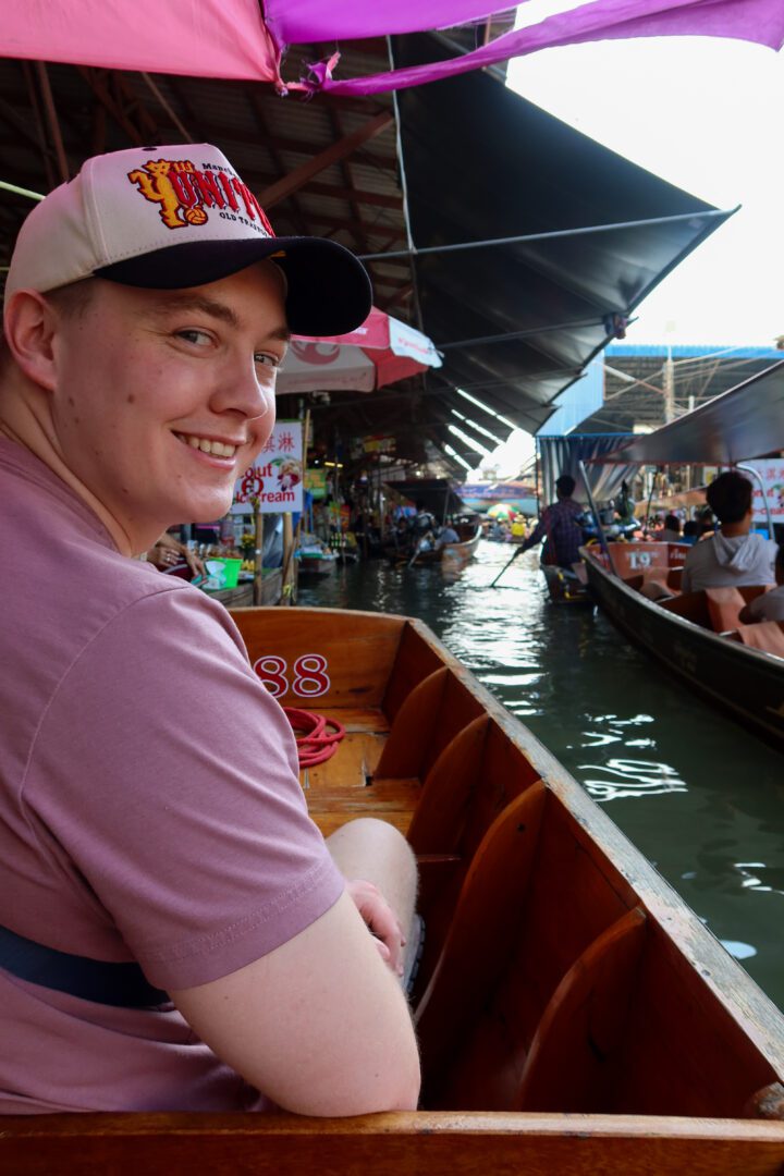 A smiling man enjoys a boat ride through Damnoen Saduak Floating Market, surrounded by a maze of longtail boats and vibrant vendor stalls lining the canal.