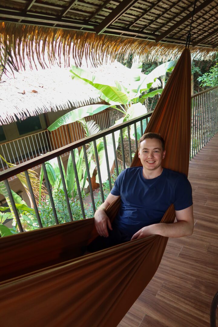 A man relaxes in a hammock on the shaded balcony of his jungle tent at Elephant Hills, surrounded by lush tropical greenery in Khao Sok National Park.