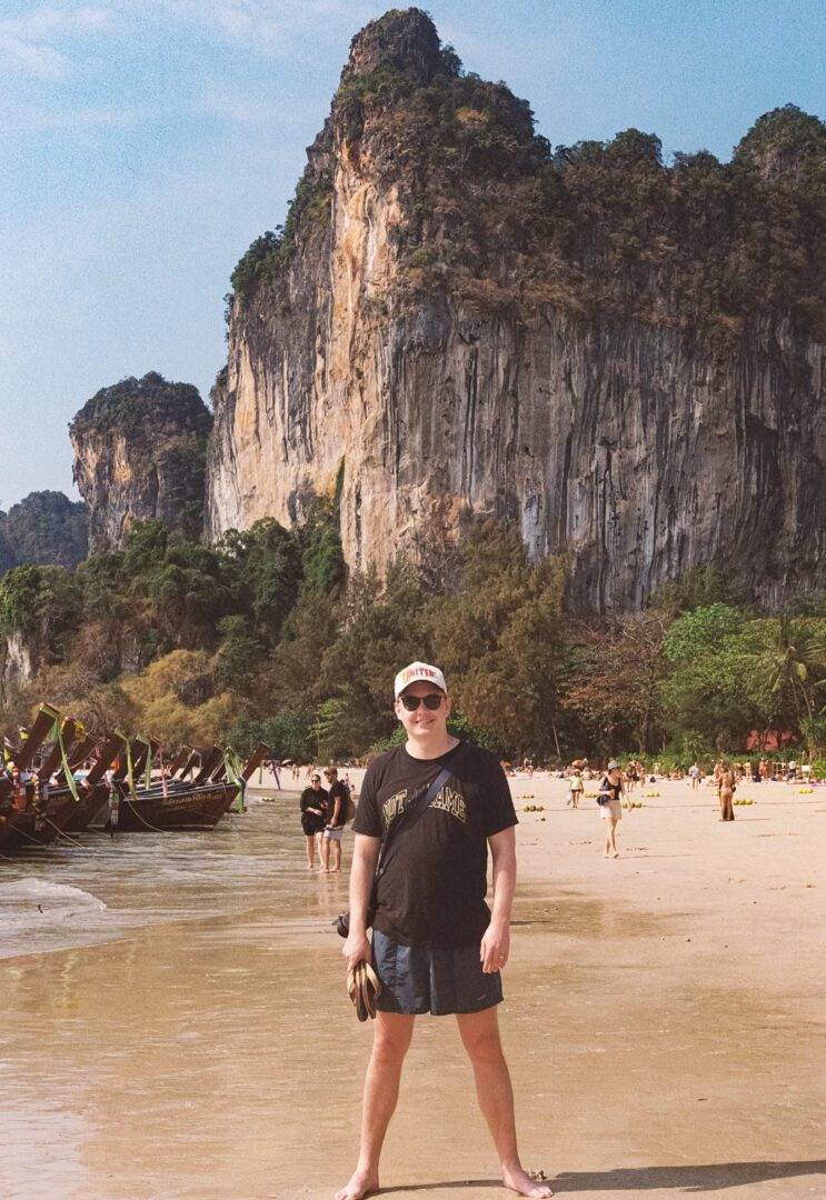 Man standing barefoot on Railay Beach in Krabi, Thailand, with longtail boats and dramatic limestone cliffs behind him.