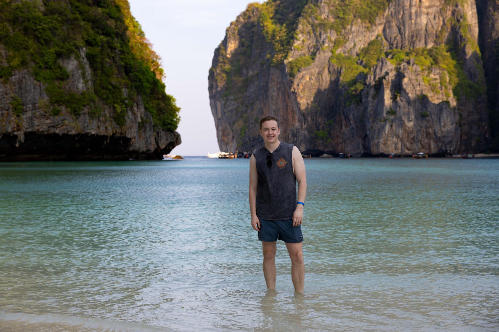 Man standing ankle-deep in clear turquoise water at Maya Bay, Koh Phi Phi, framed by towering limestone cliffs in the early morning light.