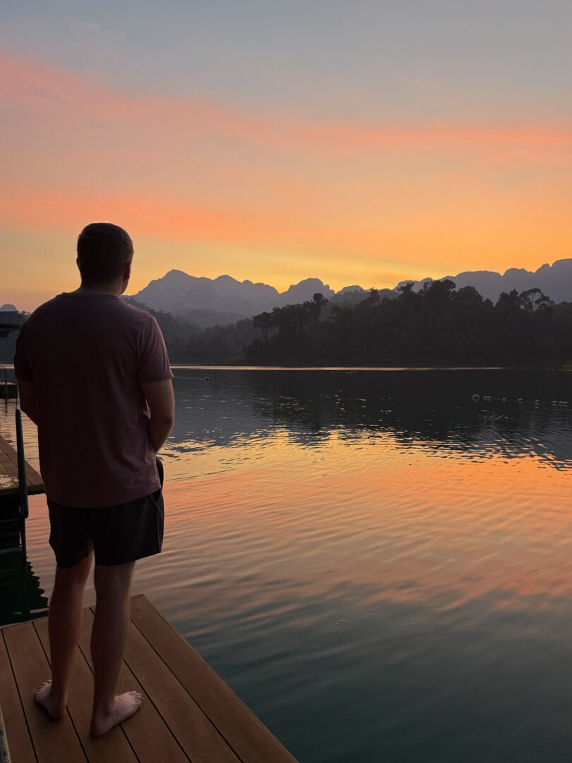 Man watching a serene sunset from a floating deck at Elephant Hills in Khao Sok National Park, with soft reflections on the lake and mountain silhouettes in the distance.