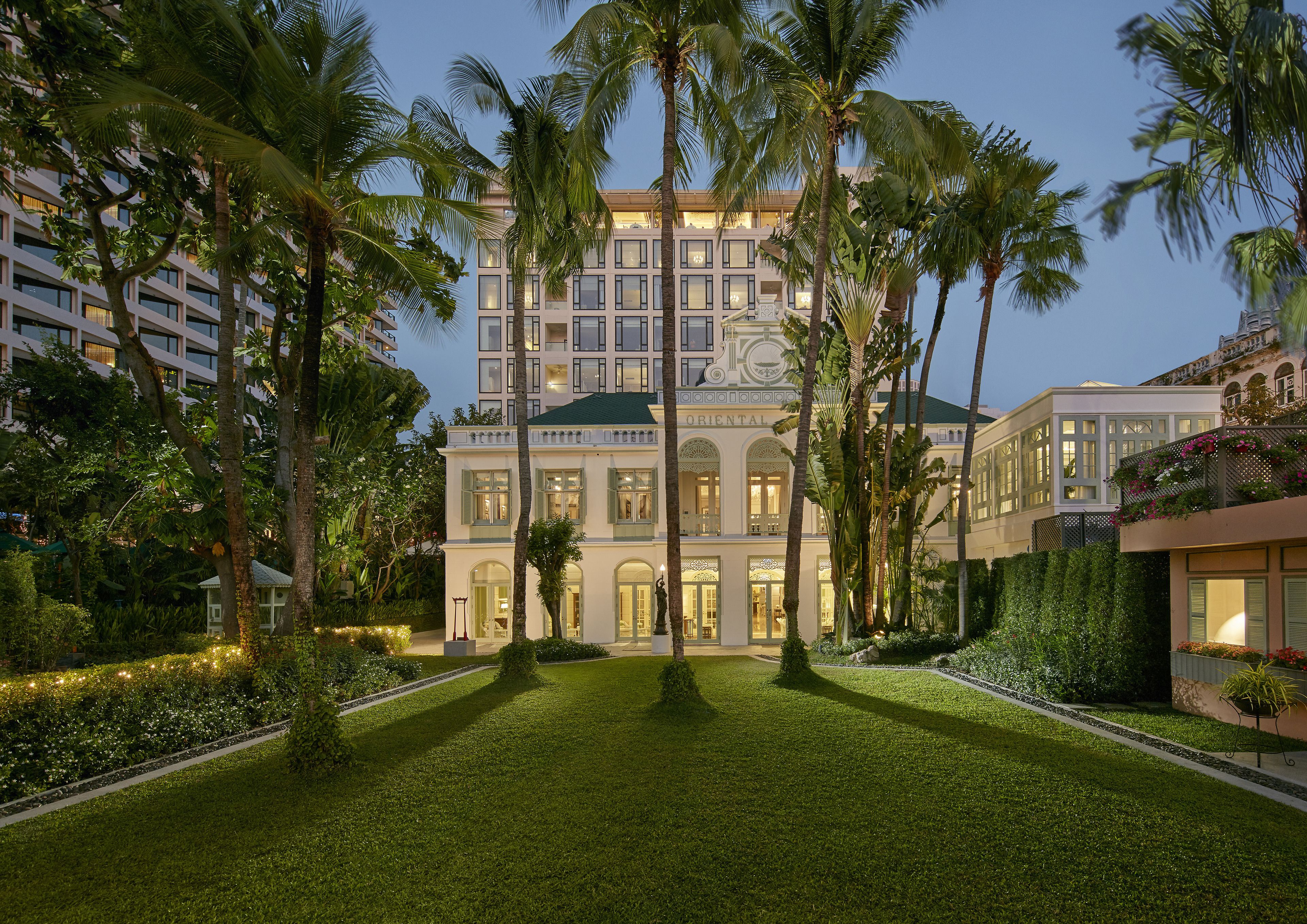 Elegant colonial-style architecture surrounded by lush gardens and tall palm trees at dusk at the Mandarin Oriental Bangkok, blending historic charm with modern luxury.