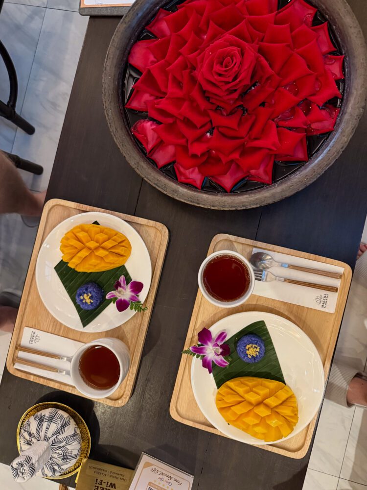 Beautifully plated mango sticky rice served with herbal tea at Makkha Spa in Chiang Mai, Thailand, presented on wooden trays with orchid garnishes and a decorative bowl of red rose petals in the background.