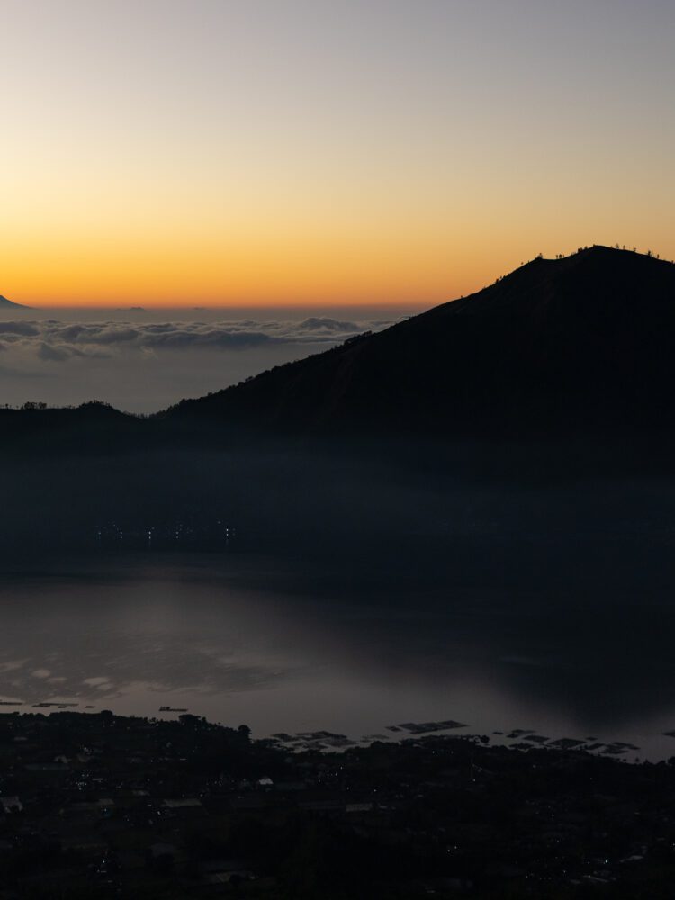 A serene view of sunrise casting an orange glow over Mount Batur and Lake Batur in Bali, Indonesia.