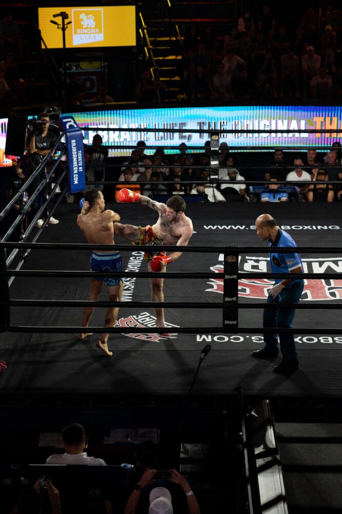 Muay Thai fighter delivering a powerful head kick during a live match in Thailand, with a referee watching closely and a packed crowd cheering from the stands inside a professional boxing arena.