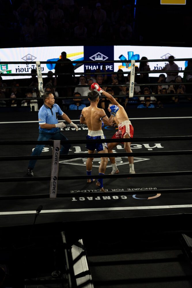 Muay Thai fighters in the middle of an intense match at a professional boxing ring in Thailand, with one fighter landing a knockout punch as the referee and audience watch closely.