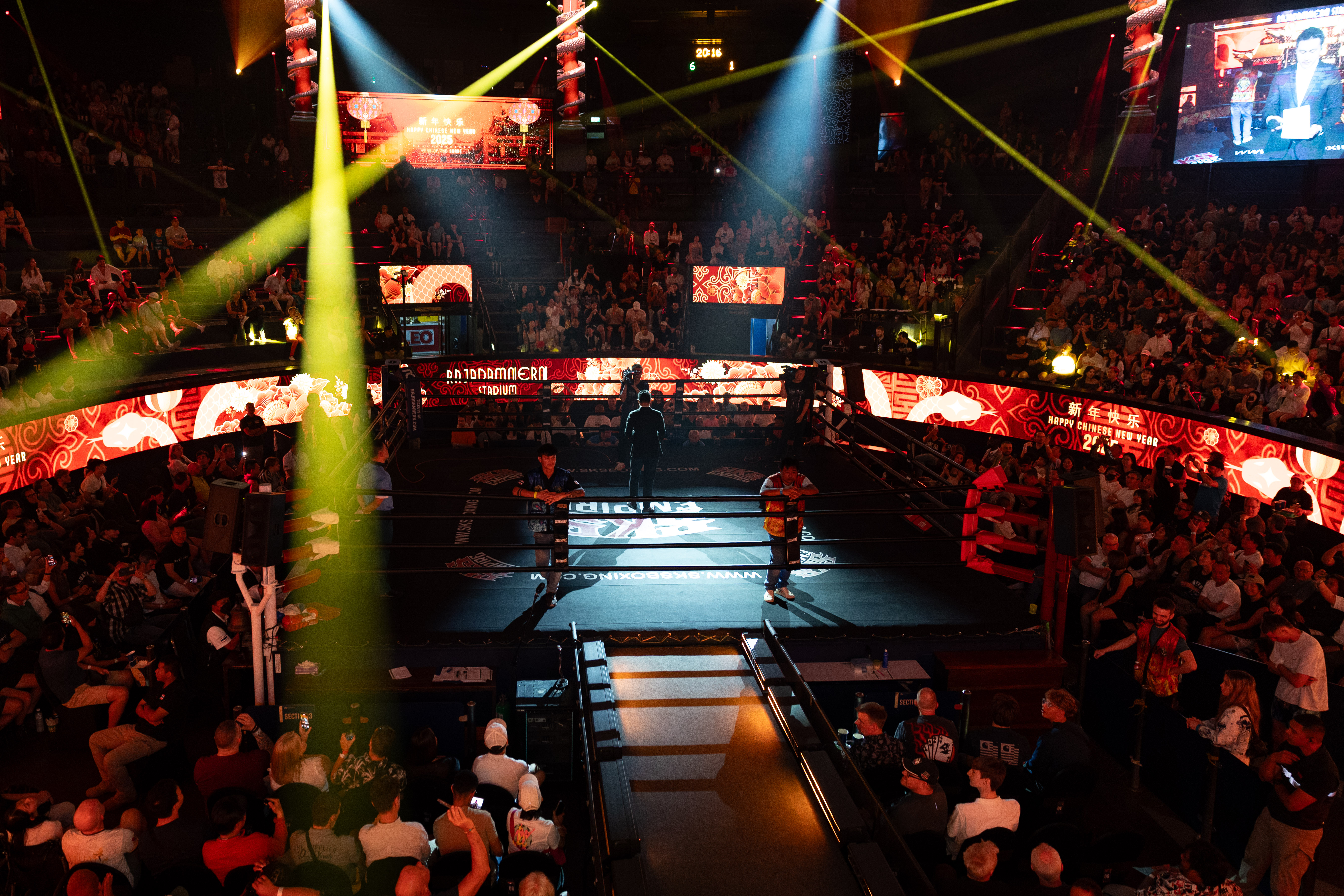 Muay Thai fighters prepare to face off under dramatic lighting at Rajadamnern Stadium in Bangkok, surrounded by a packed crowd and vibrant red decor celebrating Chinese New Year.