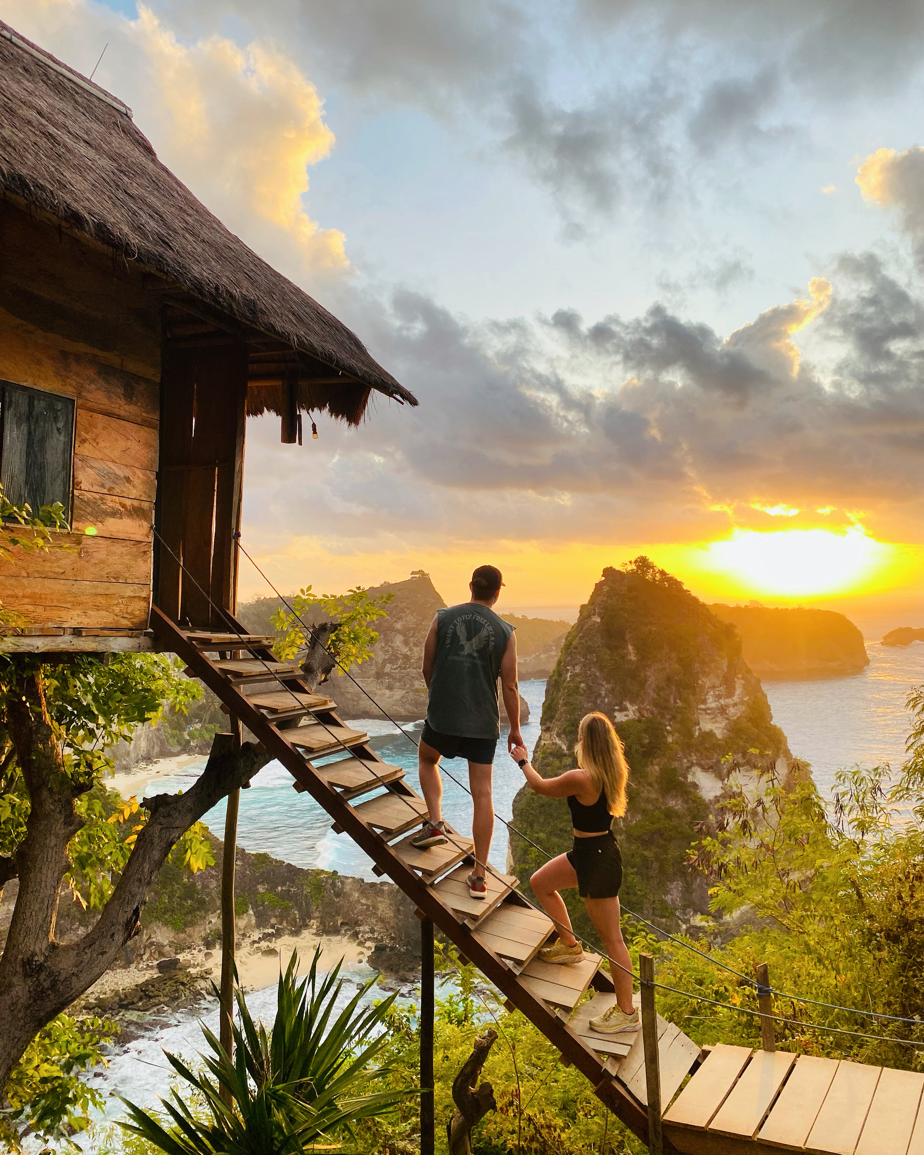 Couple climbing the stairs to a cliffside treehouse at sunrise, overlooking the iconic rock formations of Nusa Penida’s Thousand Islands Viewpoint.