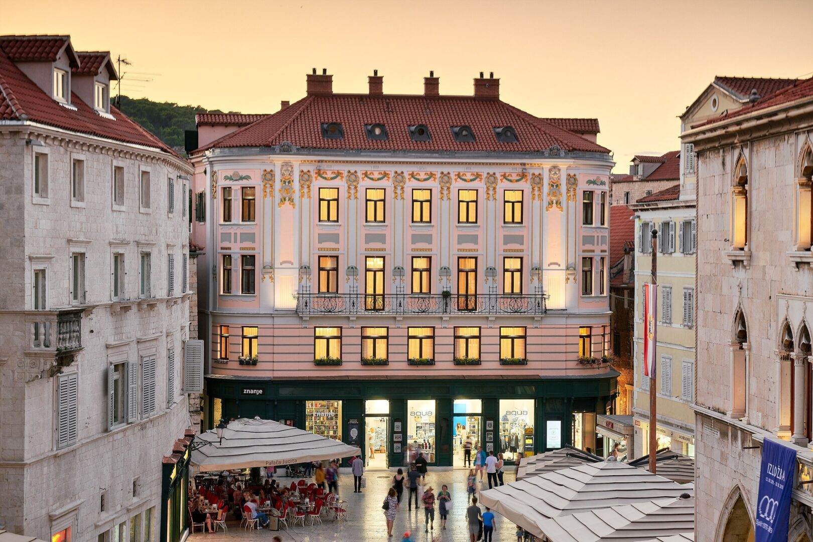 Elegant historic hotel building in Split, Croatia's main square with ornate pastel facade, viewed at golden hour as locals and tourists stroll below.