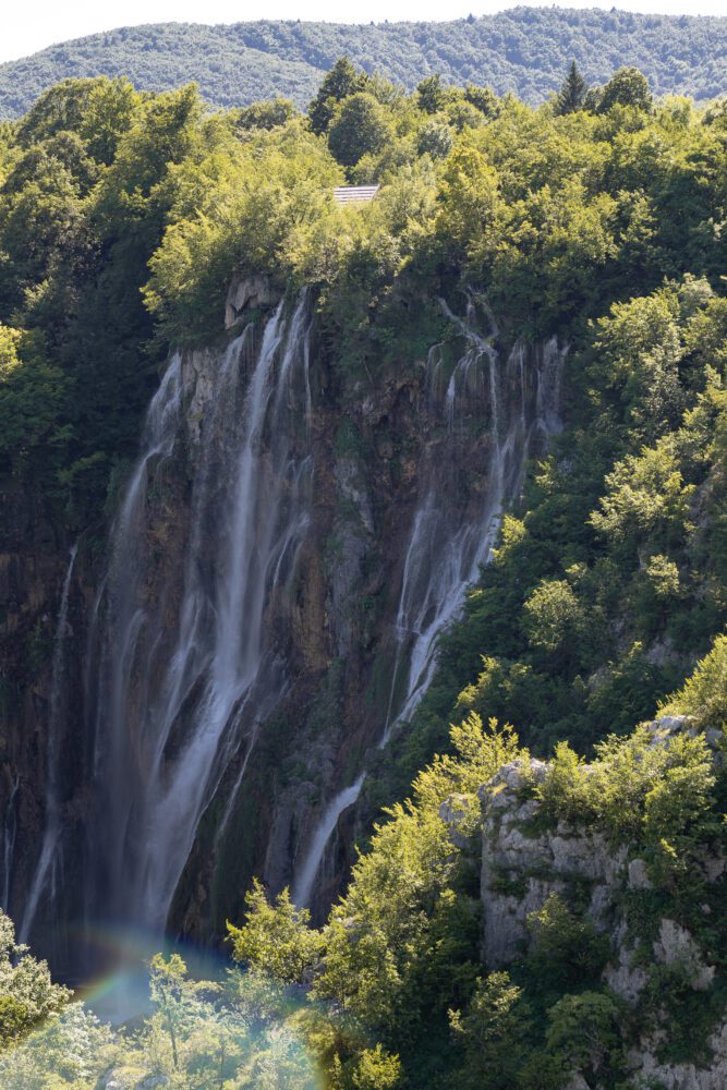 Dramatic vertical view of the Great Waterfall (Veliki Slap) at Plitvice Lakes National Park in Croatia, with powerful streams cascading down a forested cliffside—framed by lush greenery and a subtle rainbow mist near the base.
