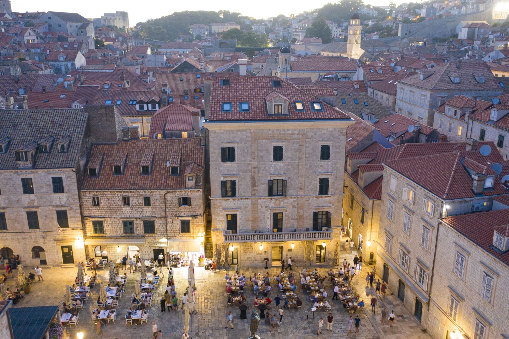 Evening view of The Pucić Palace in Dubrovnik’s Old Town, with its stately stone façade overlooking a lively square filled with open-air dining and the charm of historic terracotta rooftops.
