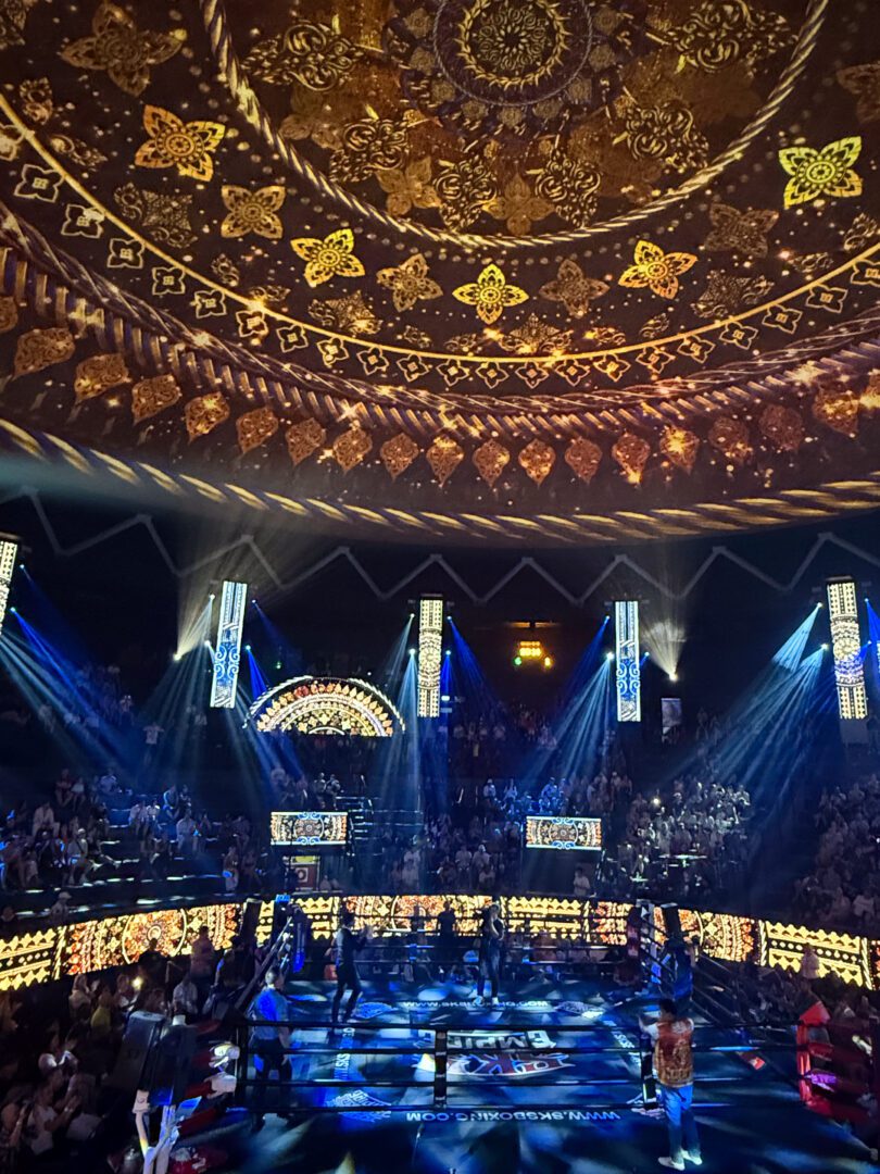 A vibrant light show fills Rajadamnern Stadium in Bangkok as performers and fighters prepare in the ring, with intricate golden patterns projected onto the dome ceiling above a packed crowd.
