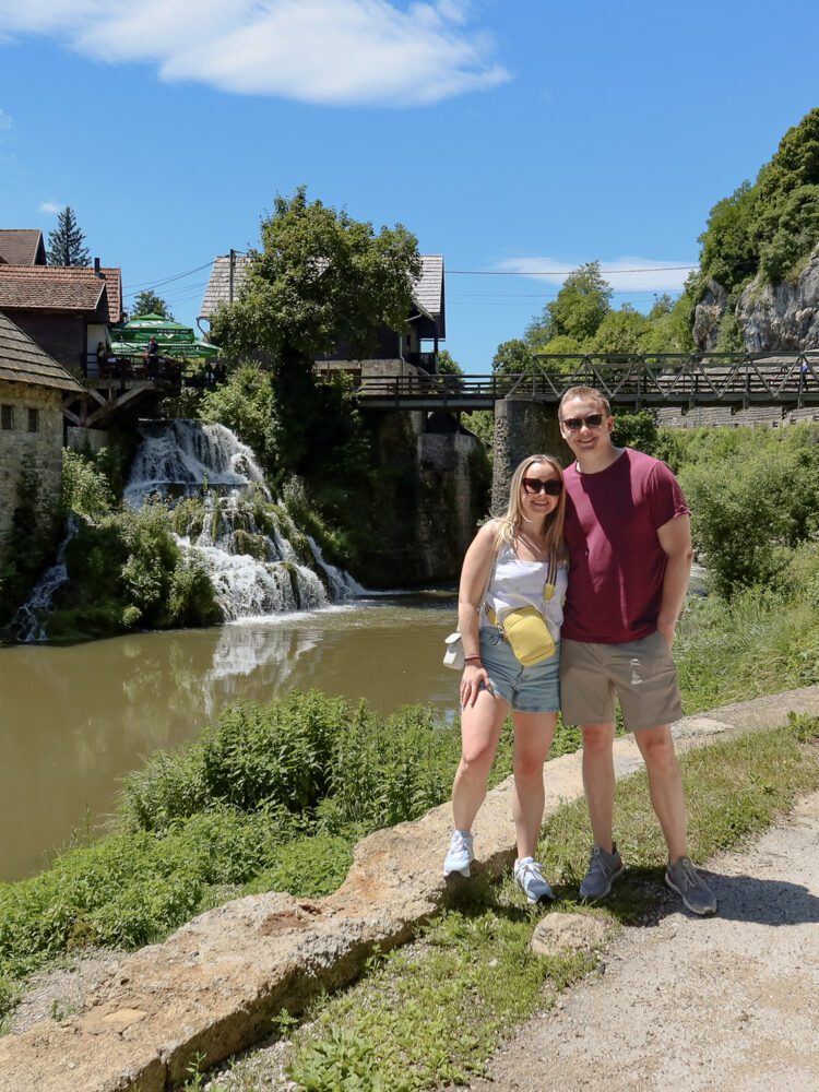 Couple standing in front of a charming waterfall and traditional wooden houses in the village of Rastoke, Croatia—known for its scenic riverside views, historic mills, and fairytale-like ambiance.