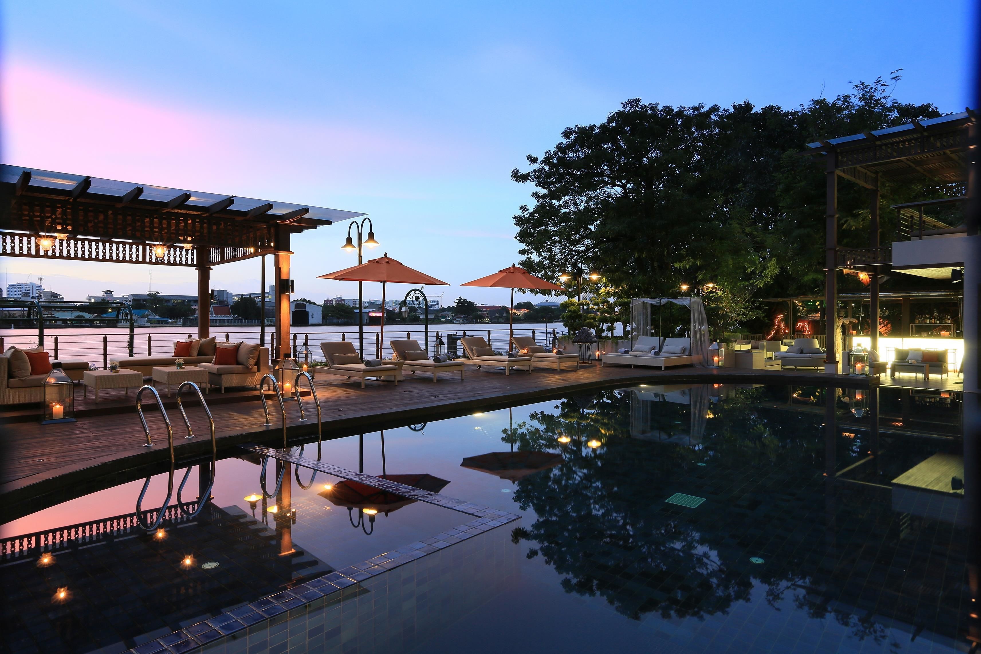 Riverside pool deck at Riva Surya Bangkok, lit by soft evening glow and city reflections, with plush loungers and umbrellas overlooking the Chao Phraya River.