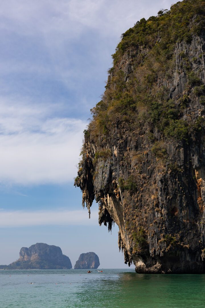 Dramatic limestone cliff formation rises above the emerald waters near Rayavadee Resort, with kayakers paddling beneath its overhang and distant islets dotting the horizon.