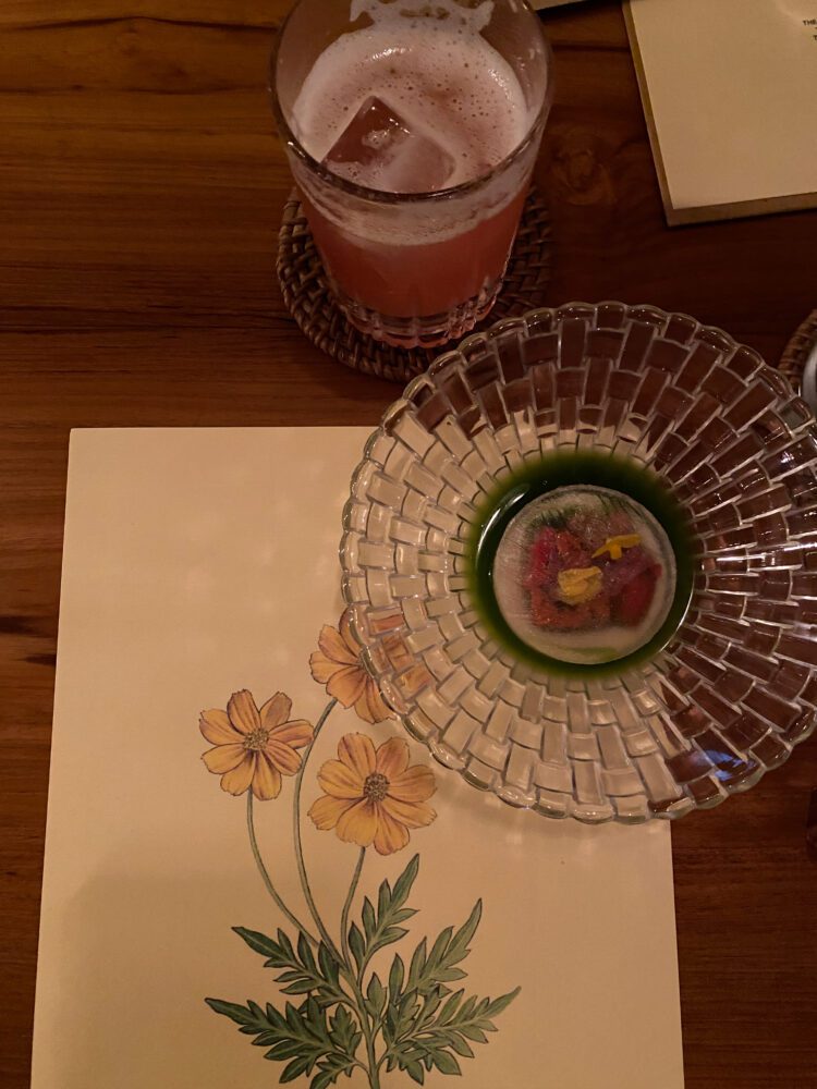 Artistic welcome course at Room 4 Dessert in Ubud, featuring a delicate floral bite and vibrant cocktail on a botanical placemat.