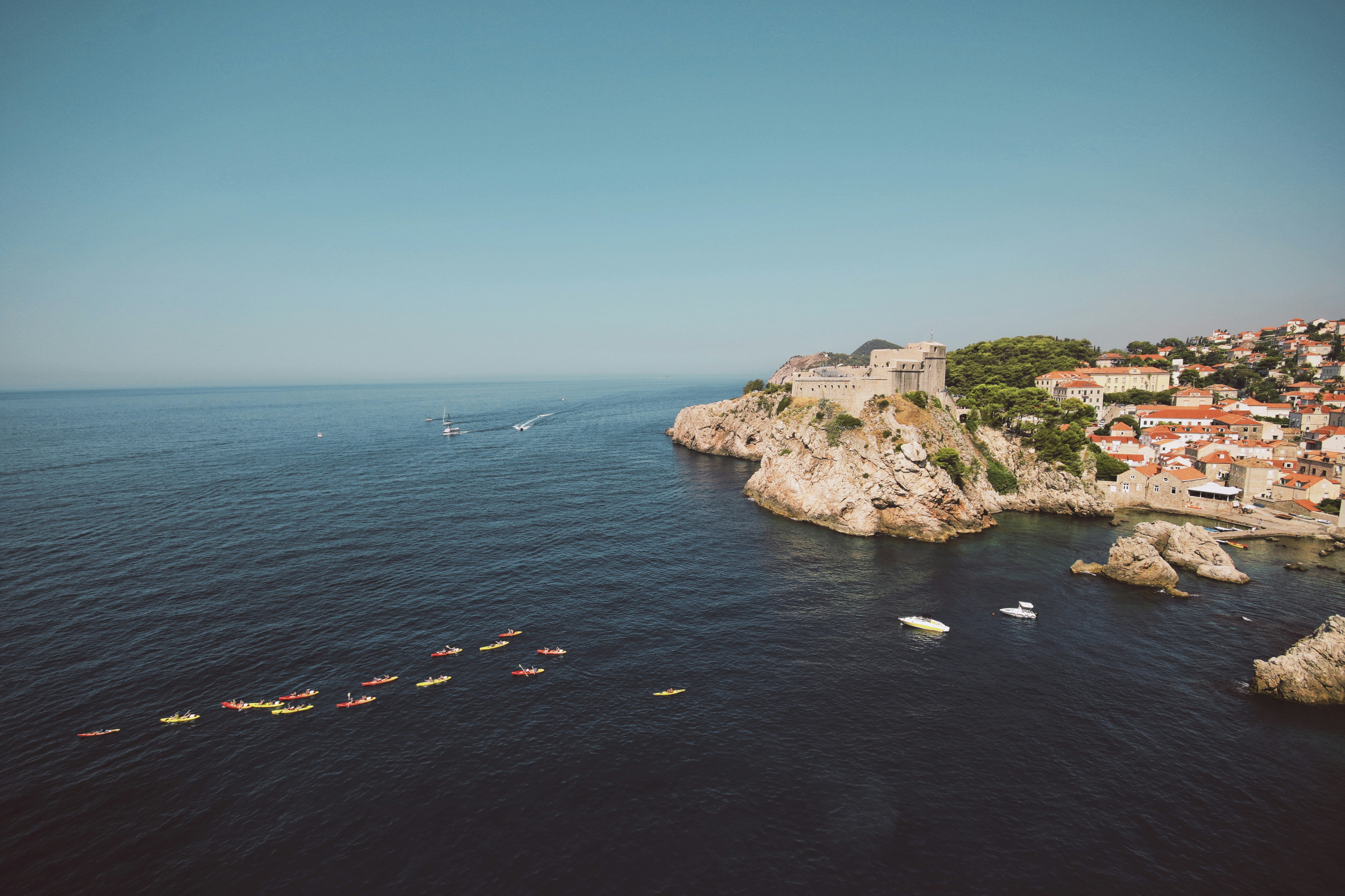 Colorful sea kayaks paddle along the Adriatic coast beneath Dubrovnik’s historic Fort Lovrijenac, with the Old Town and rocky shoreline in view.
