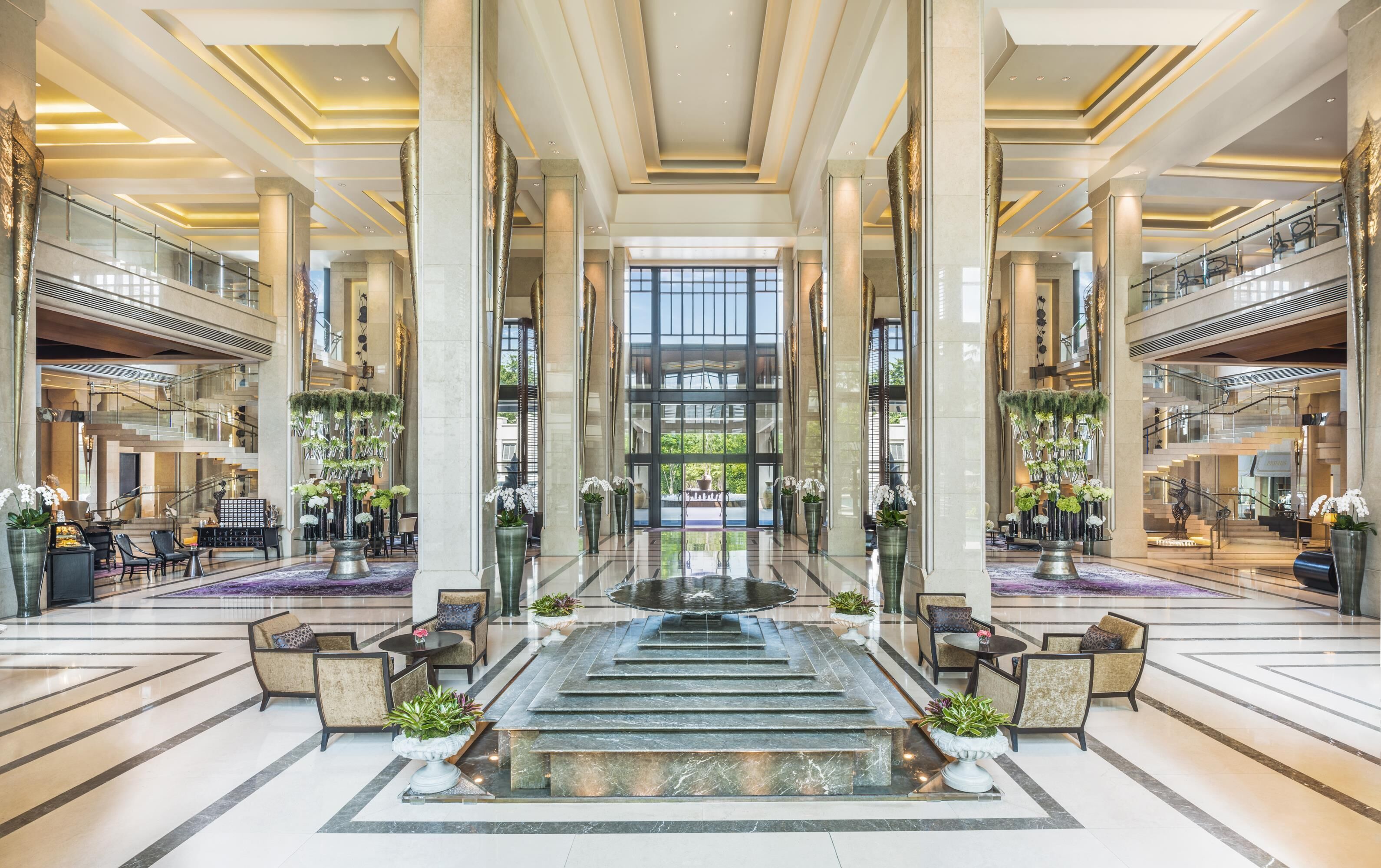 Grand lobby of Siam Kempinski Hotel Bangkok, showcasing soaring ceilings, gleaming marble floors, and a dramatic central water feature surrounded by elegant seating and towering floral arrangements.