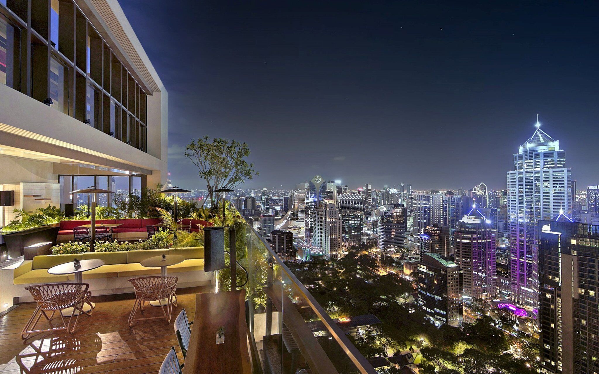 The rooftop terrace of Kimpton Maa-Lai Bangkok offers a sweeping view of the glittering Siam skyline, with cozy seating surrounded by lush greenery and the city lights sparkling below.