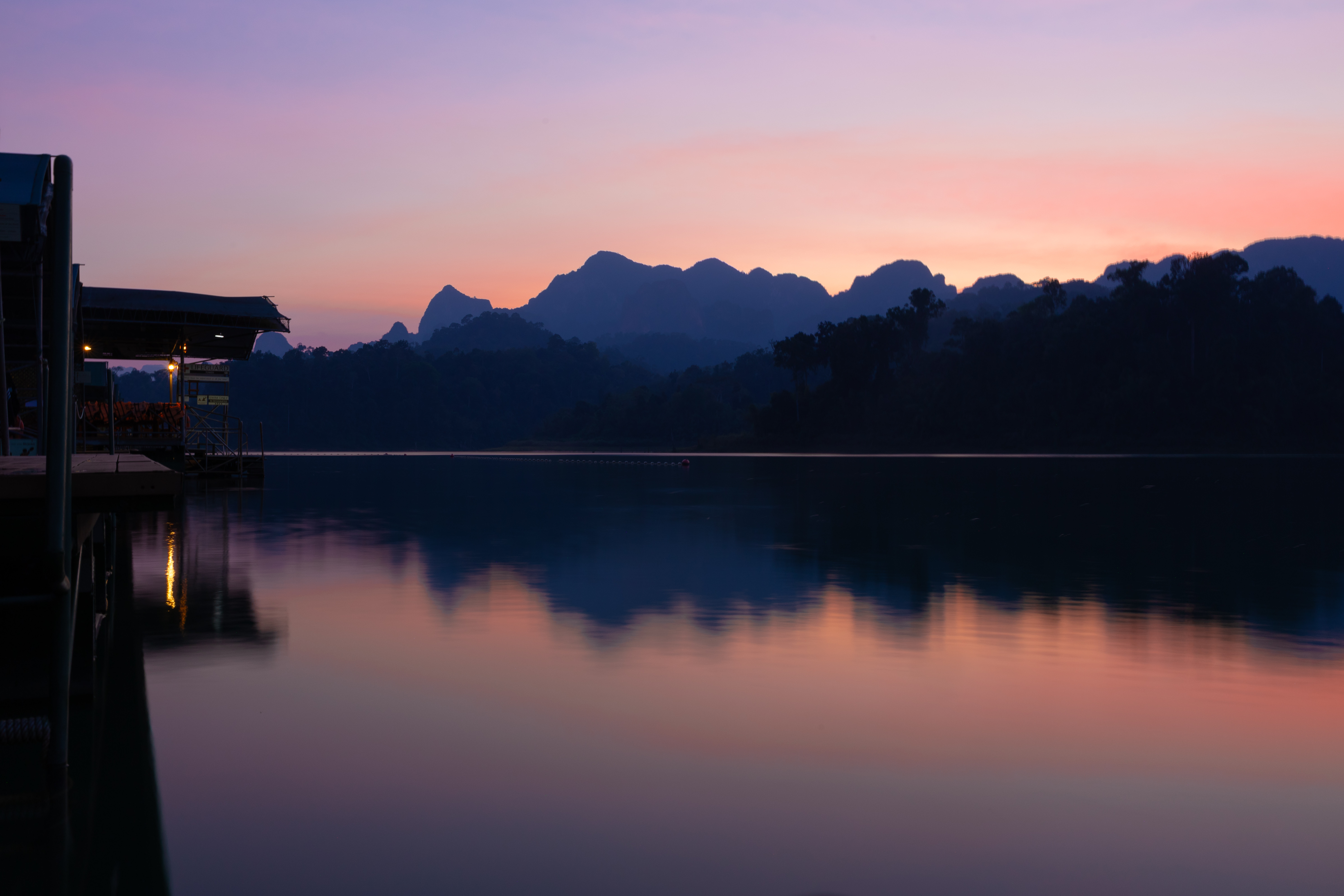 A vivid sunset casts soft pink and orange hues across the still waters of Cheow Lan Lake, silhouetting the floating tents and jagged peaks of Khao Sok National Park.
