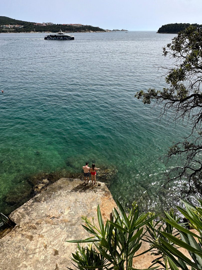 Couple standing on a stone platform over the clear turquoise waters of the Adriatic, preparing for a swim.