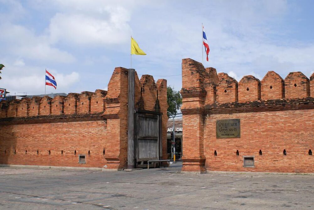 Tha Phae Gate in Chiang Mai, Thailand—an iconic historic red brick entrance to the Old City, featuring traditional battlements, Thai flags, and a large wooden door against a blue sky.